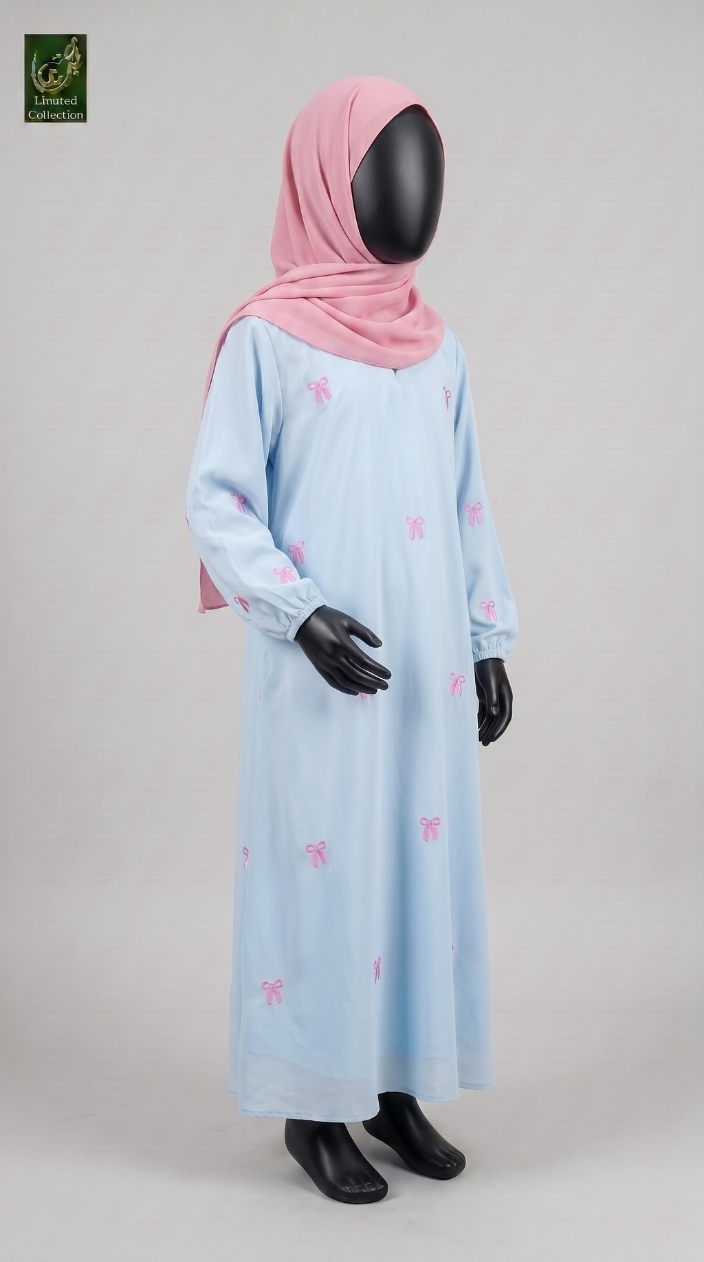 A child's long light blue abaya with small pink bow prints, styled with a pink hijab on a black mannequin against a light gray studio background.