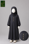 A child's long black abaya dress with a matching hijab, displayed on a white mannequin against a gray studio background. The dress features a buttoned chest detail and gathered cuffs, with a circular inset showing a close-up of the button design.