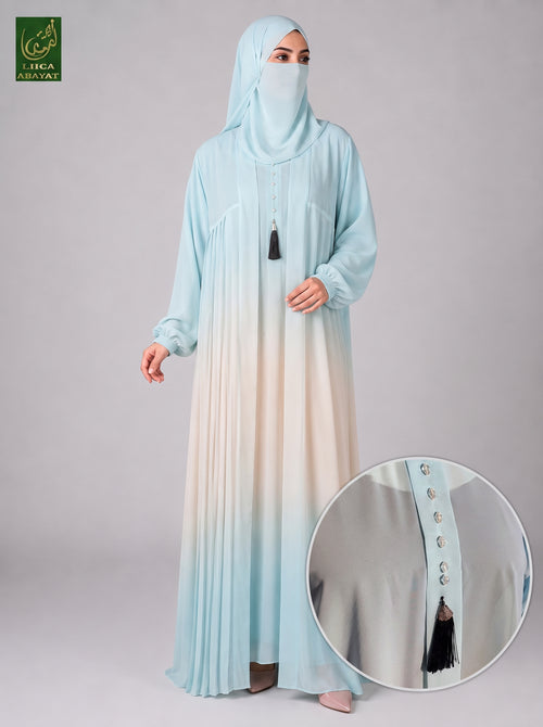 A woman modeling a light blue to cream gradient abaya with a matching hijab, shown against a neutral gray studio background. A circular inset highlights the buttoned front and tassel detail of the abaya.