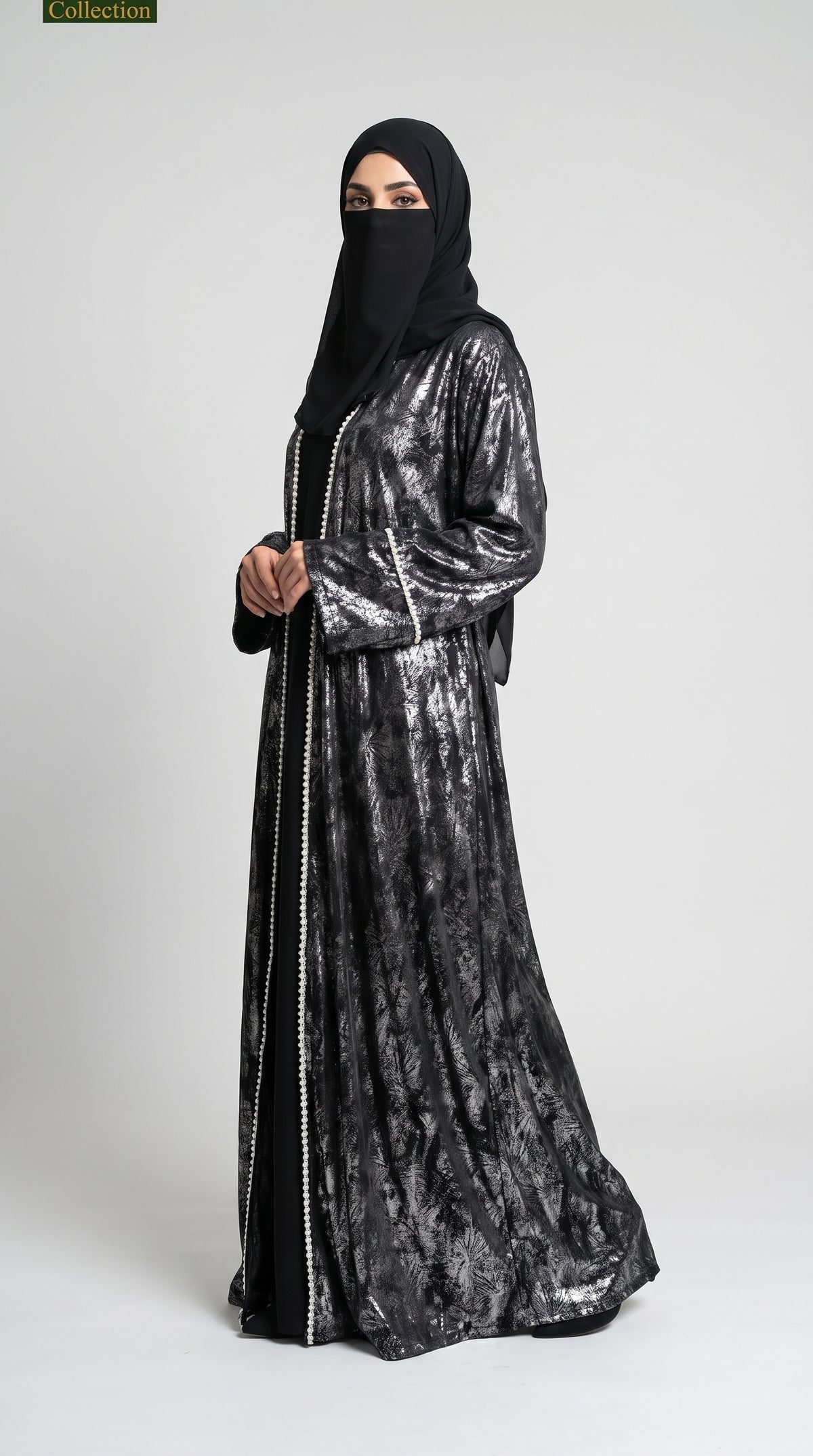 A woman wearing a long black abaya with a metallic silver pattern and gold embroidered borders, paired with a black hijab, standing against a light gray studio background.