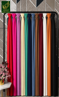 A display of multiple chiffon hijabs drape panels hanging vertically in a row, shown in a range of colors including red, pink, cream, green, blue, brown, white, orange, and black. A small floral arrangement sits on a side table in the lower left corner.