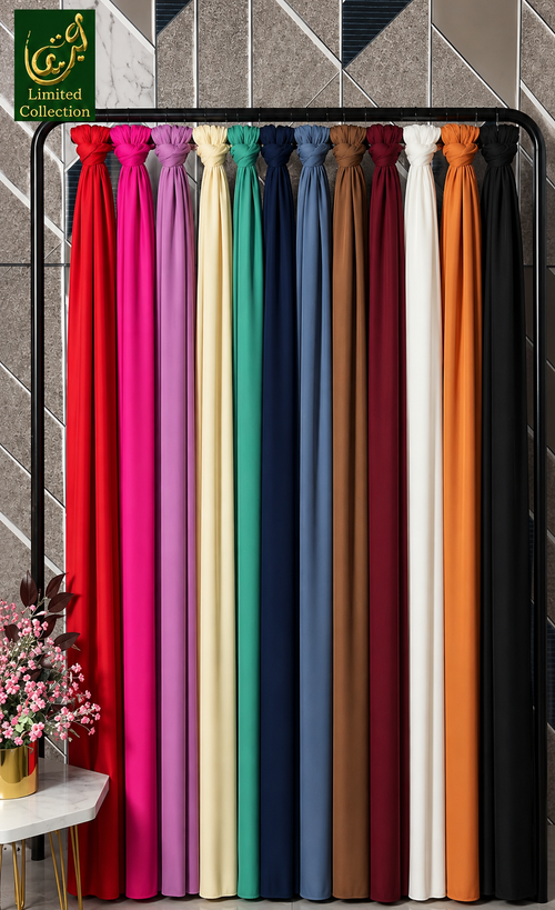 A display of multiple chiffon hijabs drape panels hanging vertically in a row, shown in a range of colors including red, pink, cream, green, blue, brown, white, orange, and black. A small floral arrangement sits on a side table in the lower left corner.