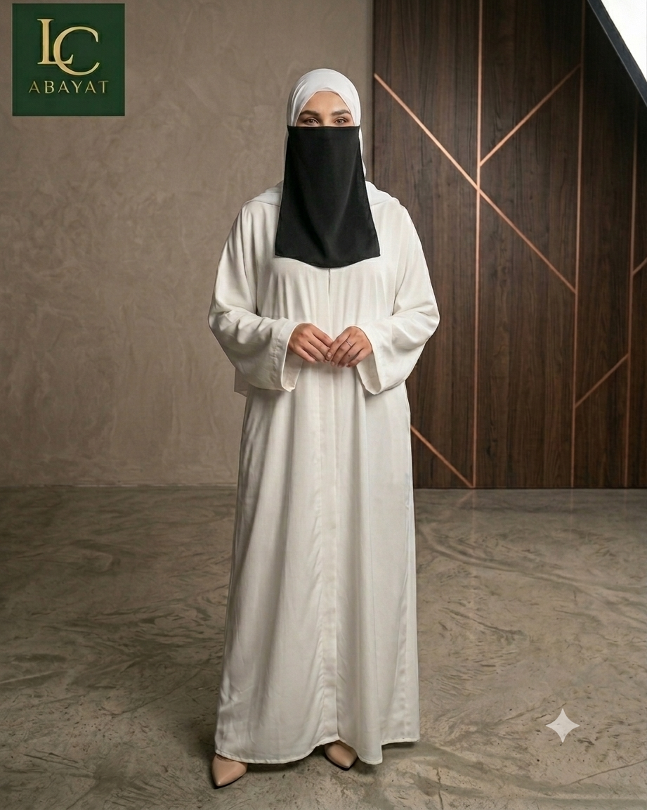 A woman wearing a long white abaya and a black face covering under a white headscarf, standing in a modern interior with a textured beige wall and dark wood paneling. The logo 'LC ABAYAT' is visible in the top left corner.