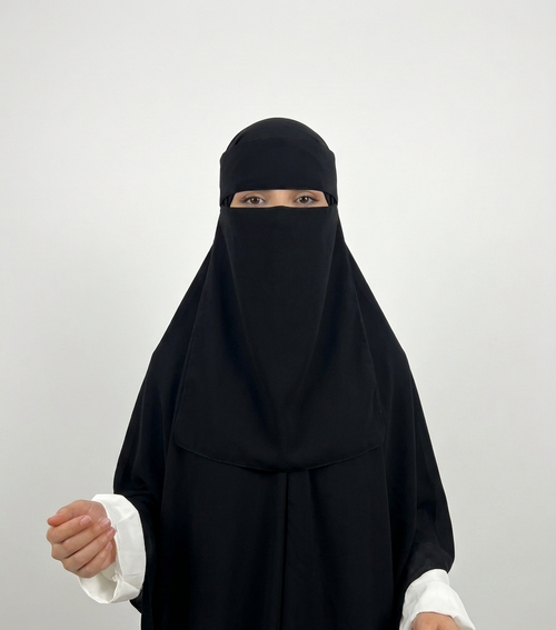 A woman wearing a black full-body modest garment with a face-covering veil and white sleeve cuffs, photographed against a plain light gray studio background.
