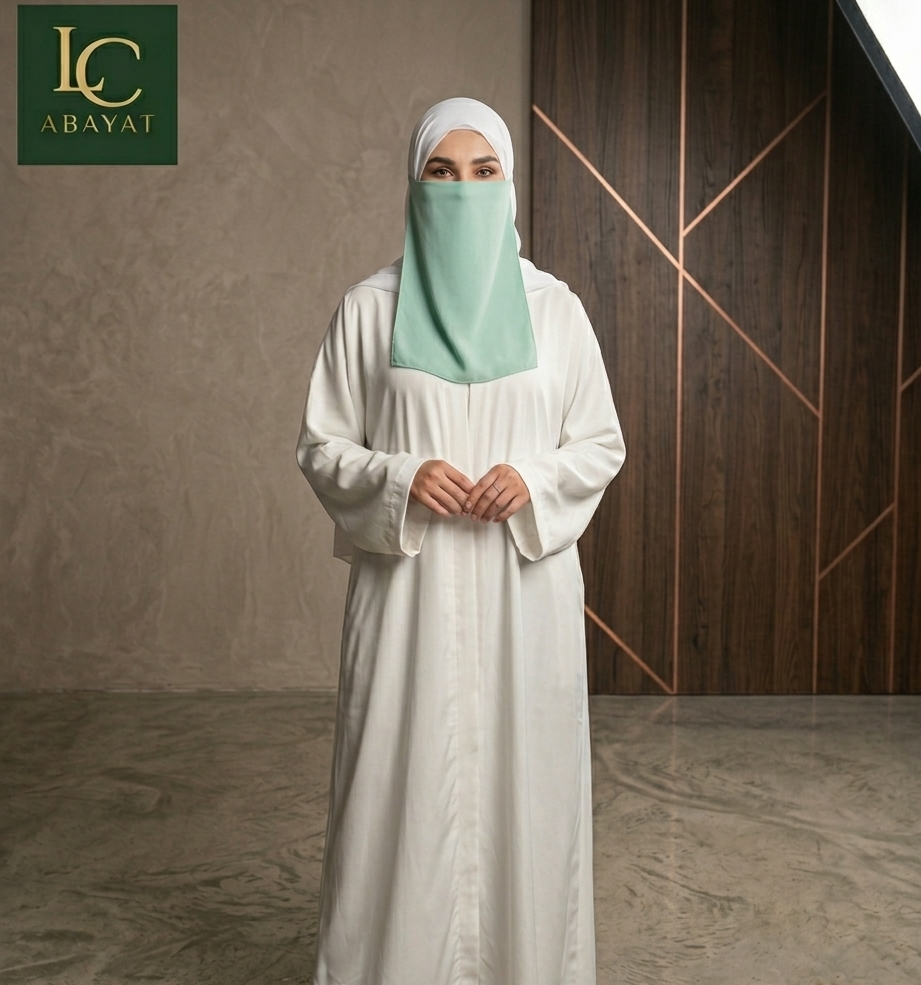 A woman wearing a white abaya and a mint green hijab stands in a modern indoor setting with a textured gray wall and dark wood paneling. The logo for 'LC Abaya' is visible in the top left corner.