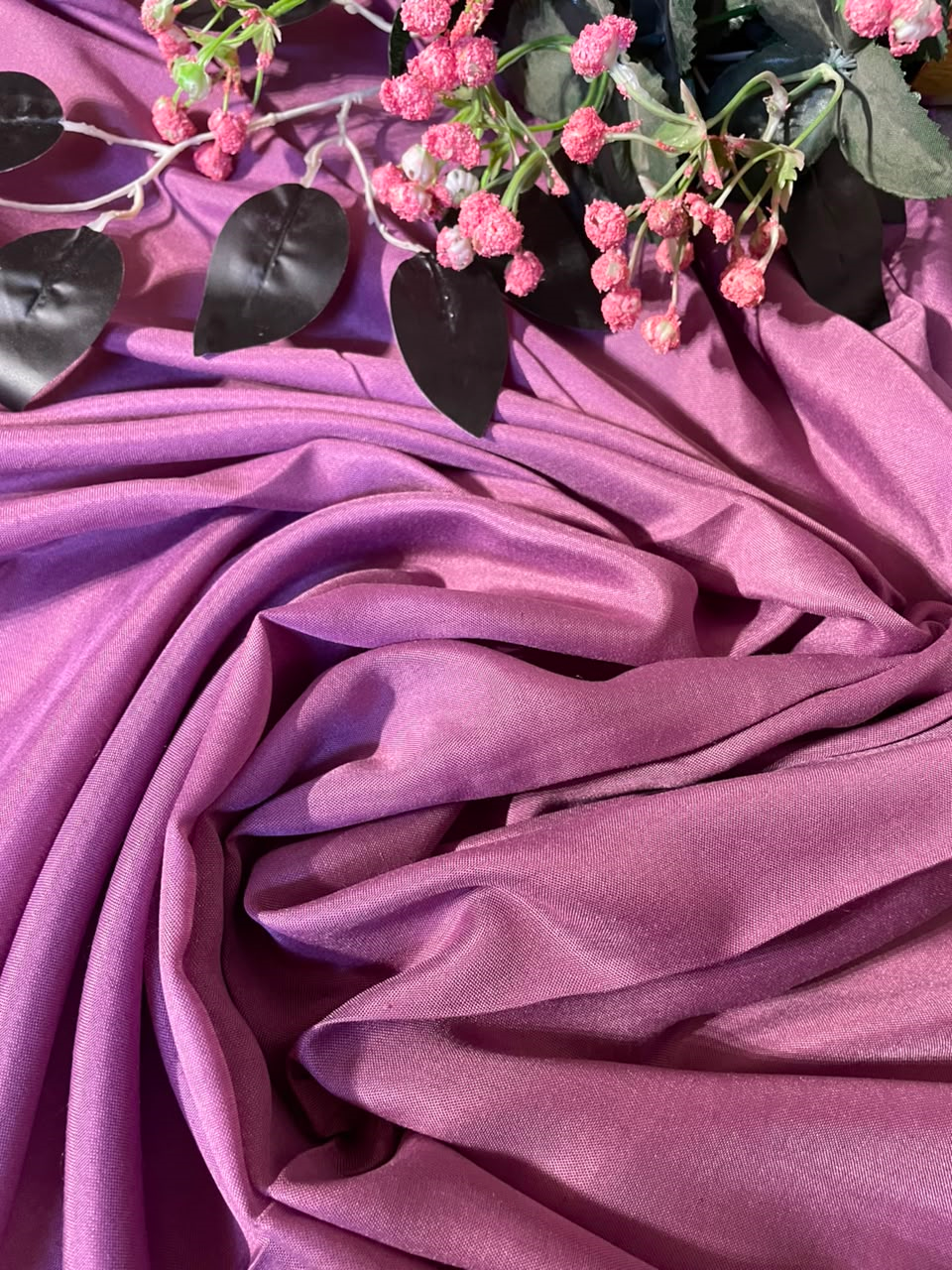 Purple draped fabric with pink flowers and dark leaves arranged on top, photographed in a styled flat-lay composition.