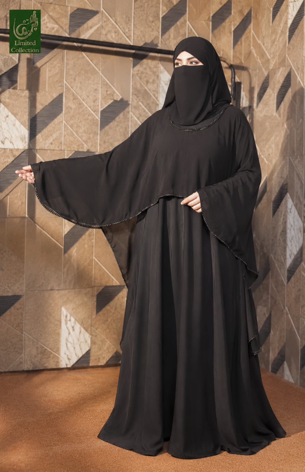 A woman wearing a full black modest Maxi Abaya with a seperate cape that has pulse work on, with a face veil and wide sleeves, standing indoors against a geometric tan wall. A green 'Ghaid Collection' logo appears in the top-left corner.