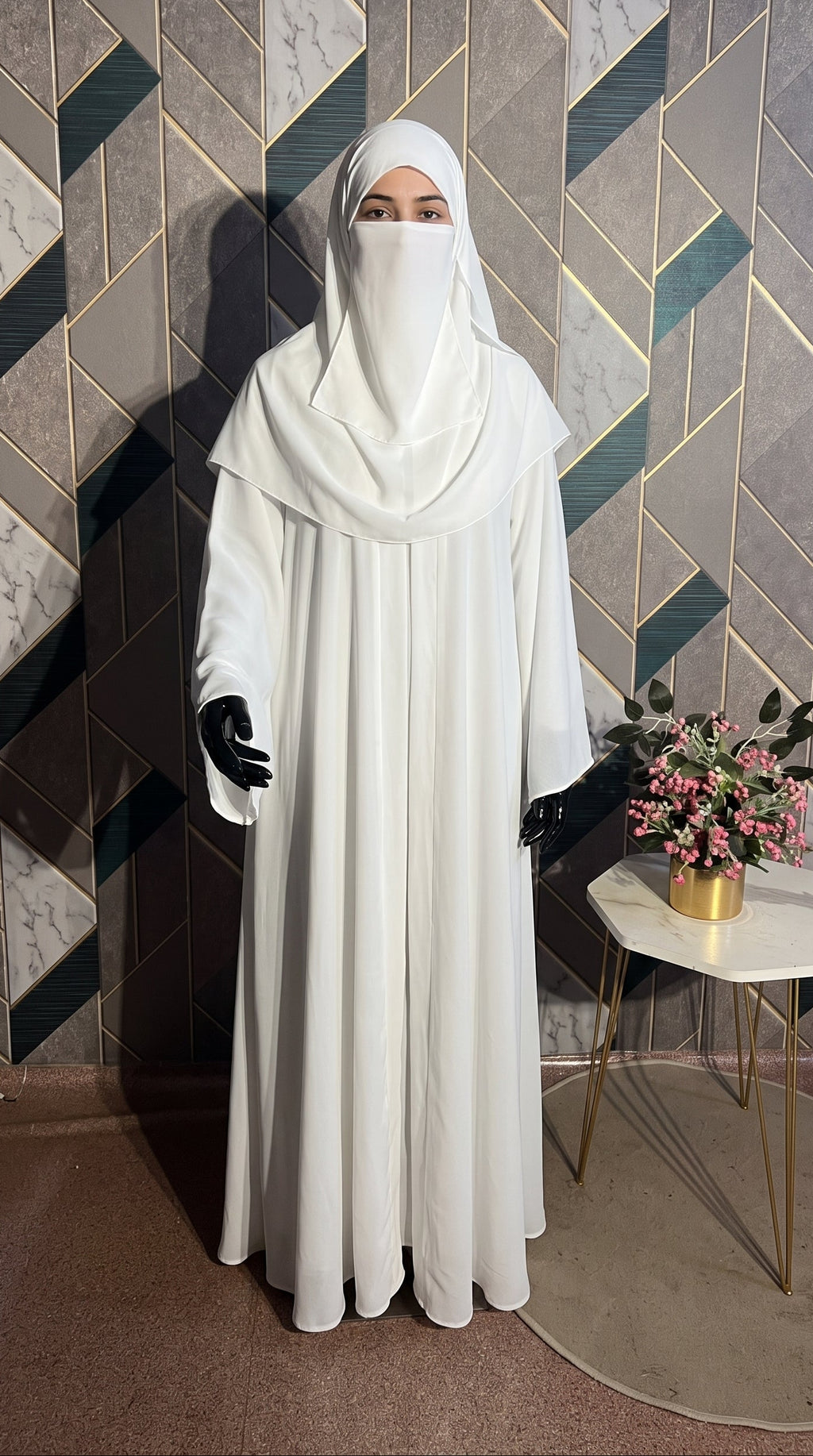 A woman wearing a full-length white front-open minimalistic modest Abaya with a matching face-covering headscarf and black gloves, standing indoors against a geometric patterned wall with a small side table and flowers beside them.