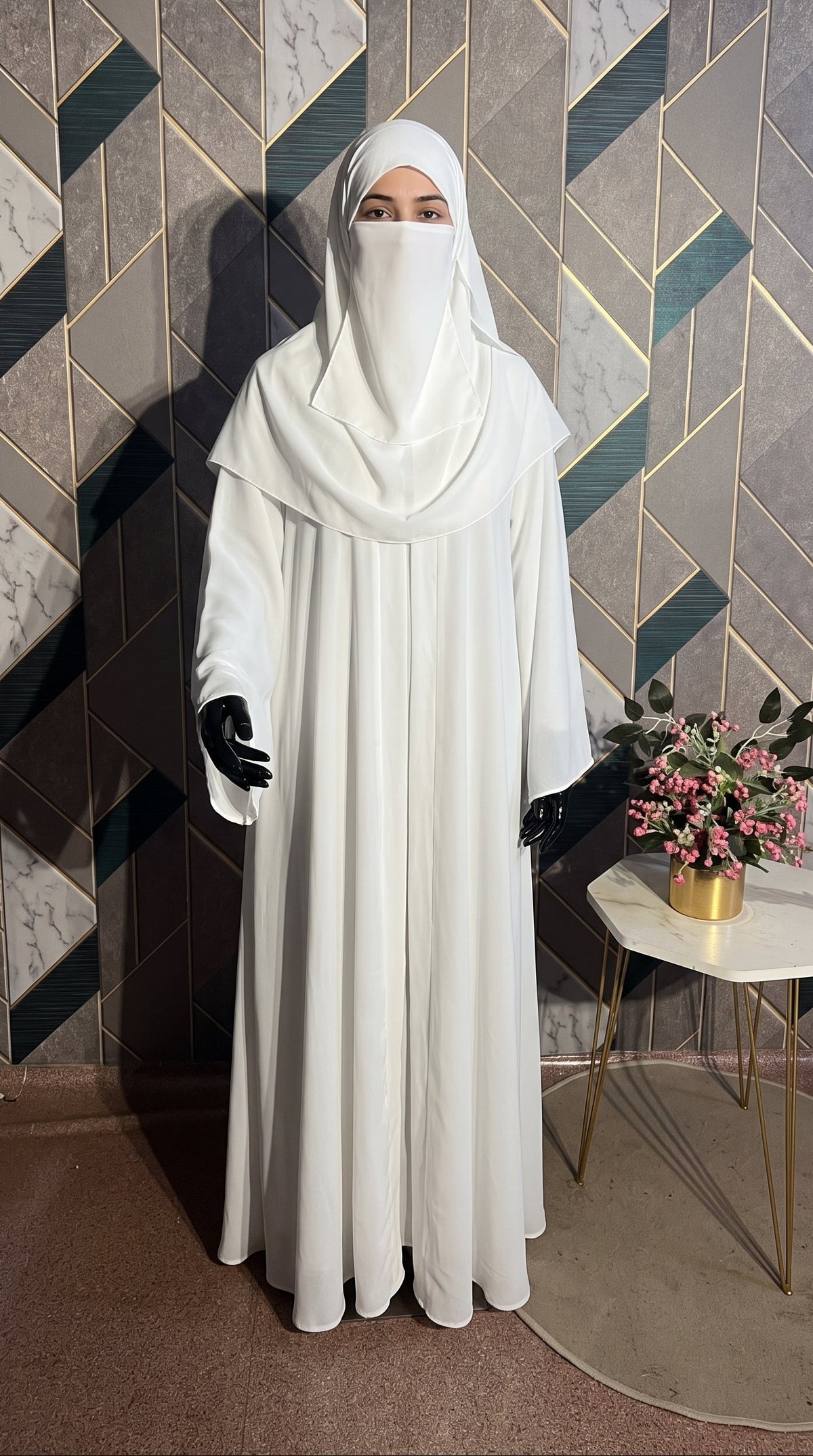A woman wearing a full-length white front-open minimalistic modest Abaya with a matching face-covering headscarf and black gloves, standing indoors against a geometric patterned wall with a small side table and flowers beside them.