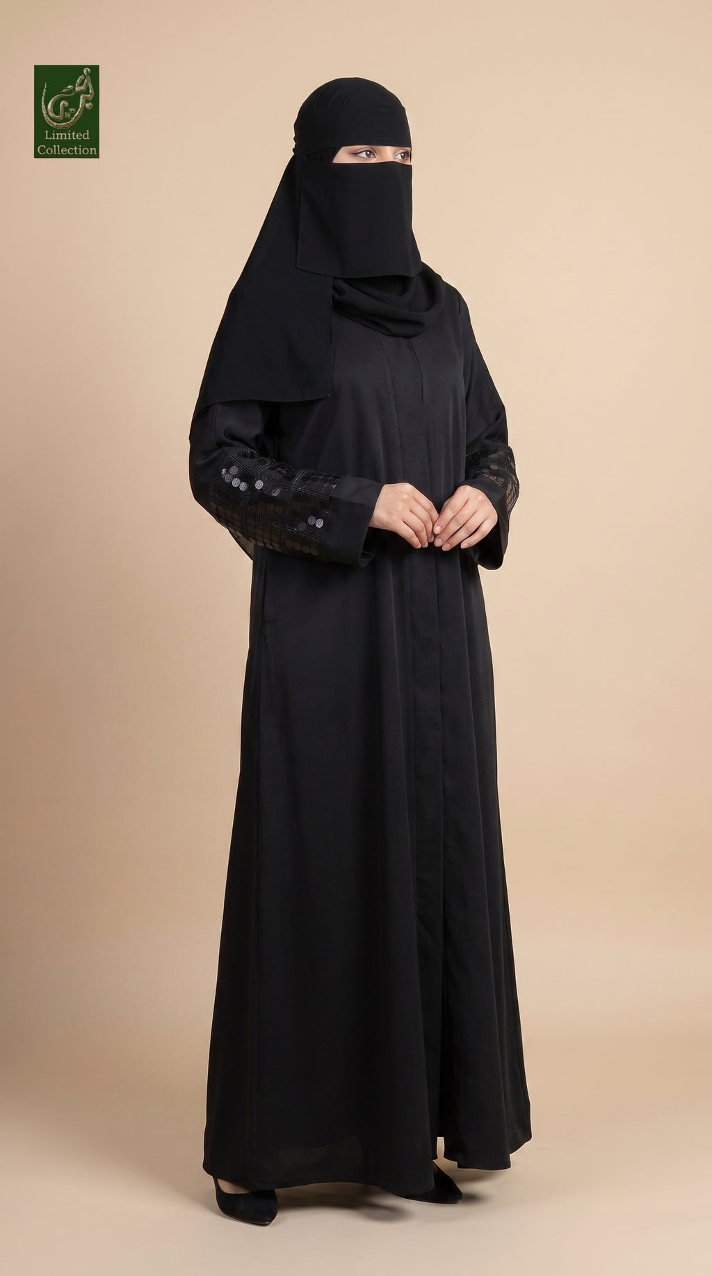 A woman wearing a full-length black abaya and matching face-covering niqab stands against a plain beige studio background. The abaya has long sleeves with subtle dark floral embroidery near the cuffs, and the model is posed with her hands clasped in front.