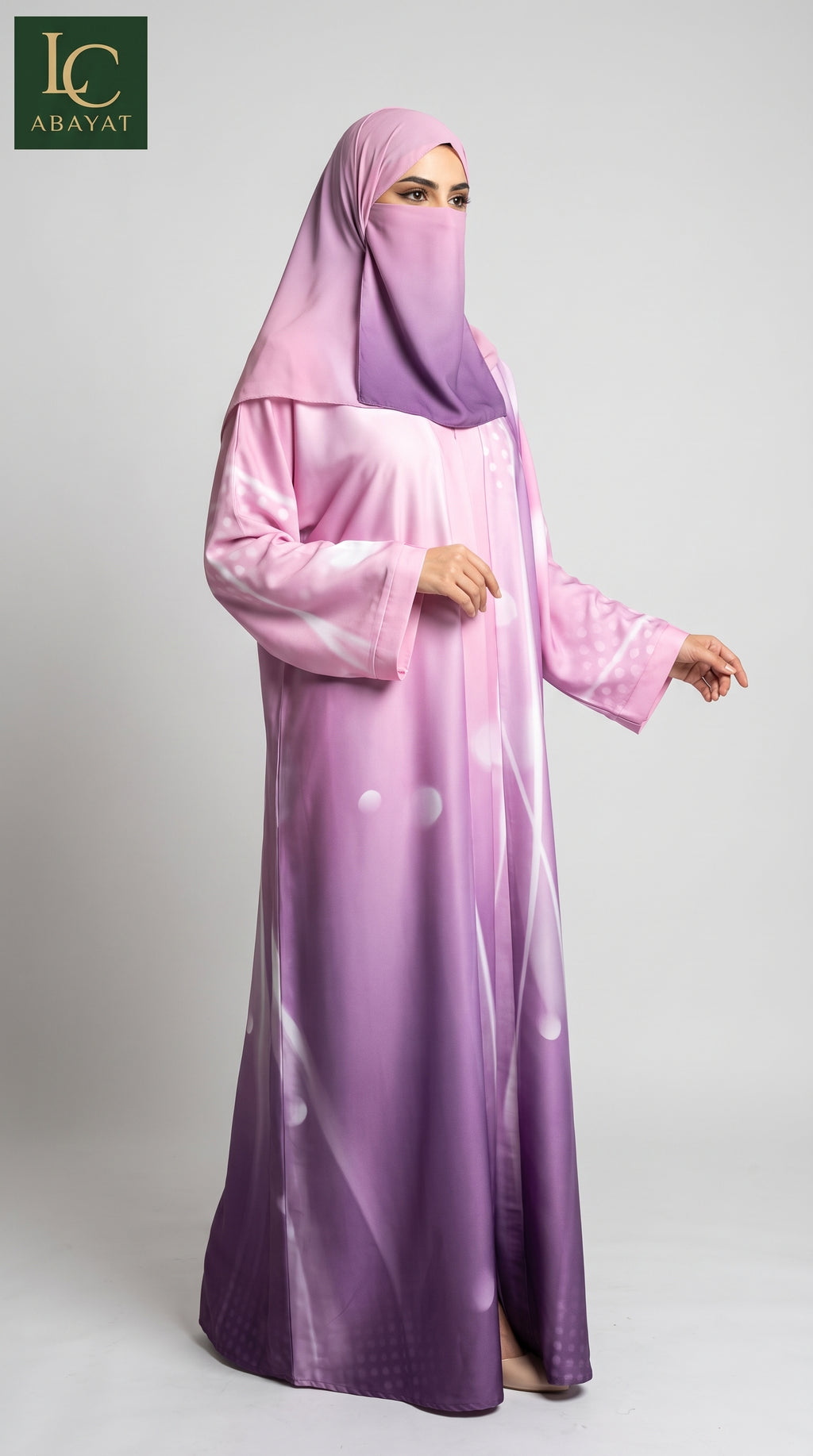 A woman wearing a long pink and purple gradient abaya with a matching hijab, standing against a light gray studio background. The abaya features a subtle white abstract pattern and a glossy finish.