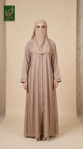 A woman wearing a long beige abaya with a matching hijab and face veil, standing against a neutral beige studio backdrop. The abaya has a subtle tonal pattern and wide sleeves.