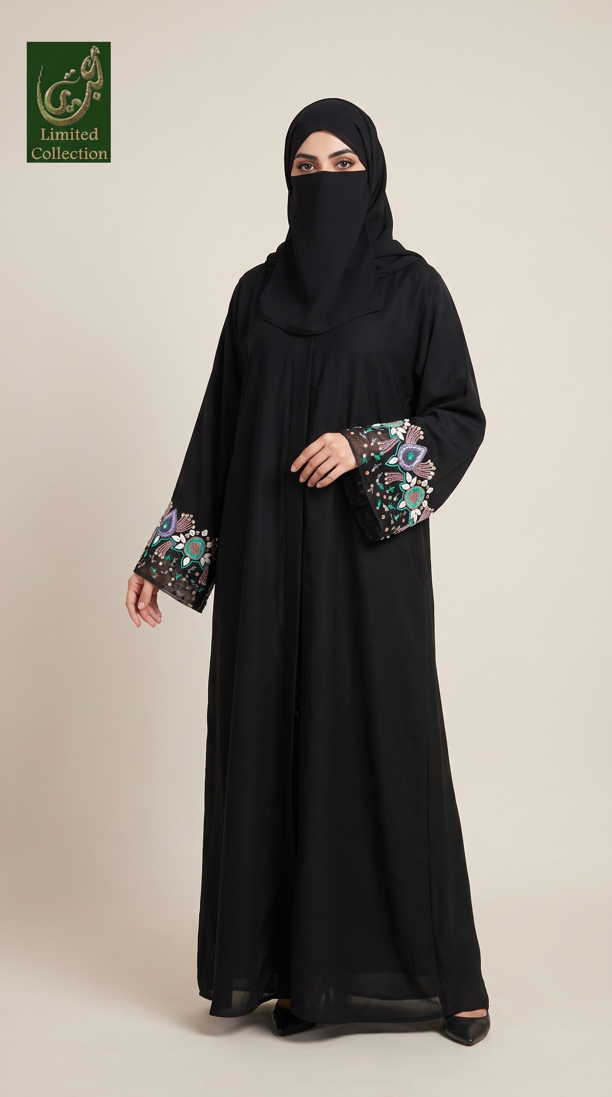A woman modeling a black abaya with a matching black hijab, featuring colorful embroidered patterns on the cuffs, set against a plain beige studio background.