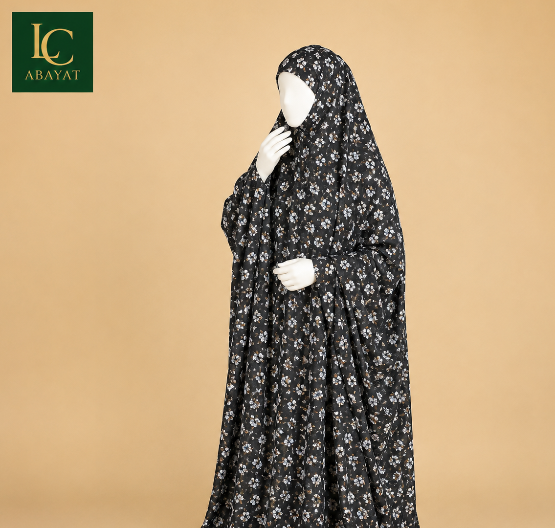 A long black modest garment with a small white floral print, displayed on a white mannequin against a warm beige studio background. The brand logo 'LC ABAYAT' appears in the top-left corner.