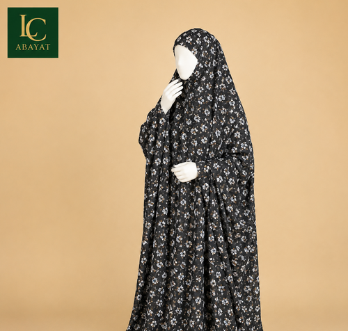 A long black modest garment with a small white floral print, displayed on a white mannequin against a warm beige studio background. The brand logo 'LC ABAYAT' appears in the top-left corner.