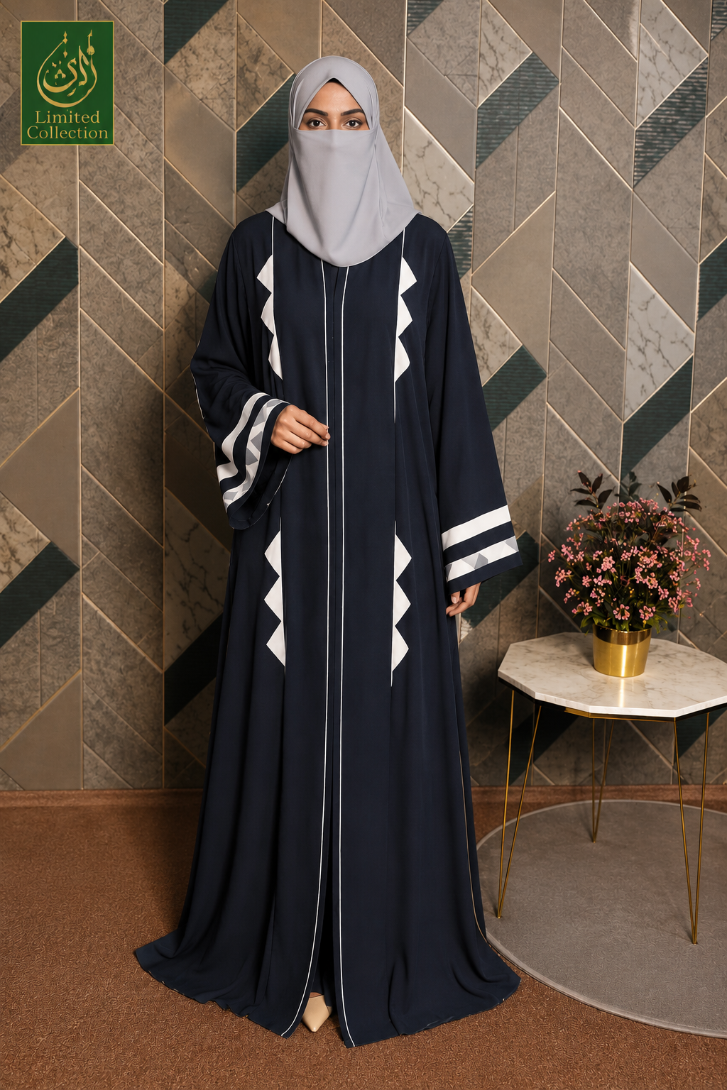 A woman wearing a long navy abaya with white geometric accents and a light gray hijab, standing in front of a geometric patterned wall. A small side table with flowers is to her right, and a green 'Limited Collection' logo appears in the top-left corner.