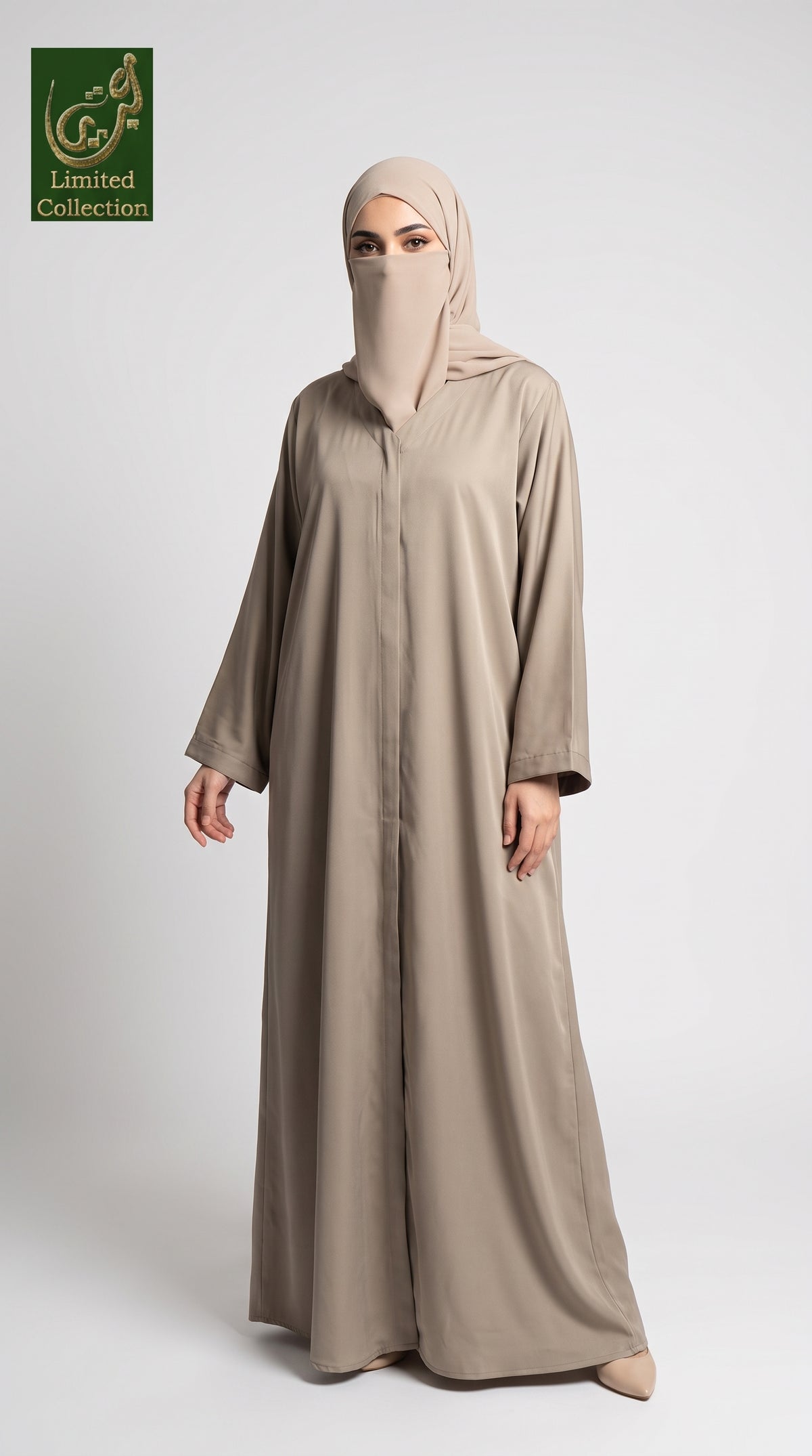 A woman modeling a long, loose-fitting abaya in a light taupe-beige color, paired with a matching hijab. She is standing against a plain light gray studio background.