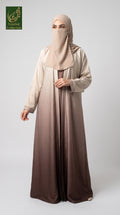 A woman modeling a modest long abaya dress with a matching hijab, featuring a smooth gradient from light beige at the top to deep brown at the bottom, set against a plain light gray background.