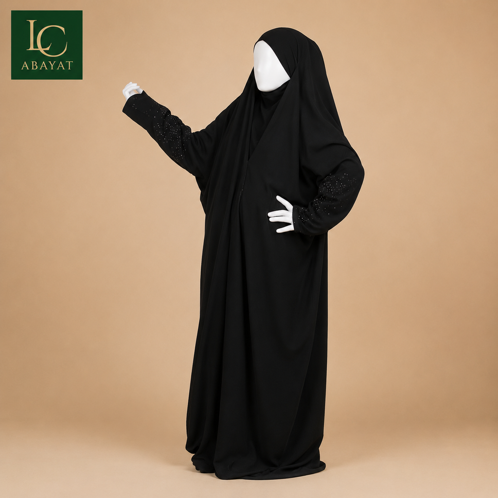 A black full-length modest garment with a hood and long sleeves, displayed on a white mannequin against a warm beige studio background. The brand logo 'IC ABAYAT' appears in the top-left corner.