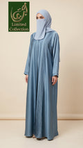 A woman wearing a light blue abaya with vertical pleats and a matching light blue hijab, standing against a beige studio background. A green 'Limited Collection' logo is visible in the top left corner.