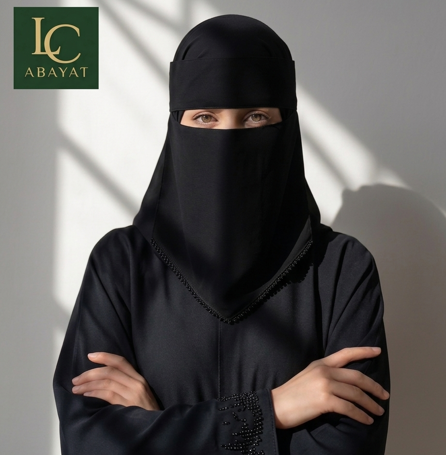 A woman wearing a black full-face niqab and matching long-sleeve abaya stands with arms crossed against a light gray wall with soft shadow patterns. The LC Abayat brand logo is visible in the top-left corner.