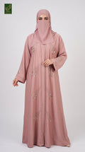 A woman wearing a dusty pink abaya and matching hijab with subtle floral embroidery, standing against a light gray studio background. A small green logo is visible in the top-left corner.
