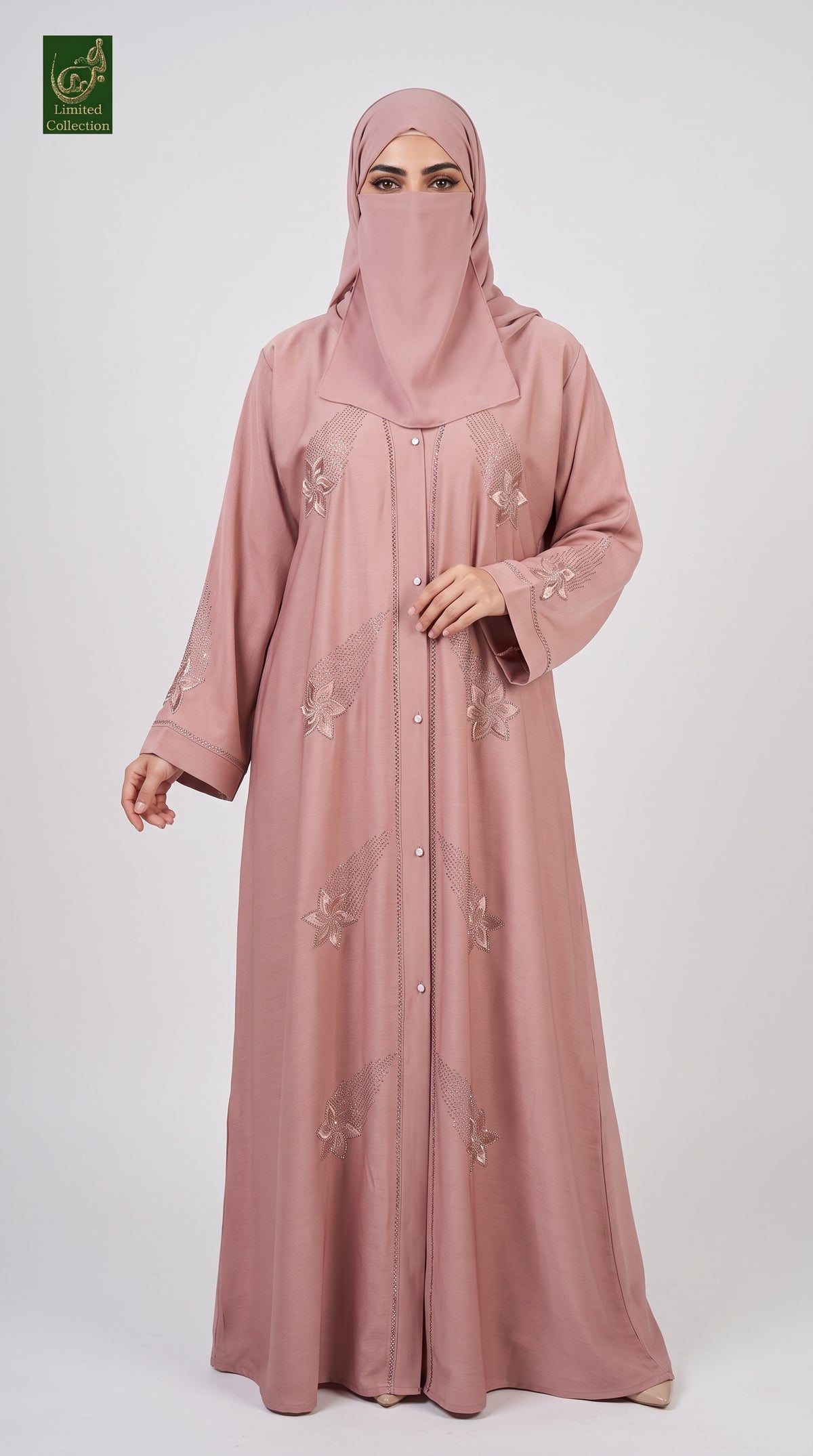 A woman wearing a dusty pink abaya and matching hijab with subtle floral embroidery, standing against a light gray studio background. A small green logo is visible in the top-left corner.
