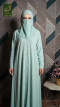 A woman wearing a light mint green long-sleeved abaya and matching hijab stands in front of a geometric tiled wall. The abaya has a subtle vertical crinkle texture and a buttoned front.