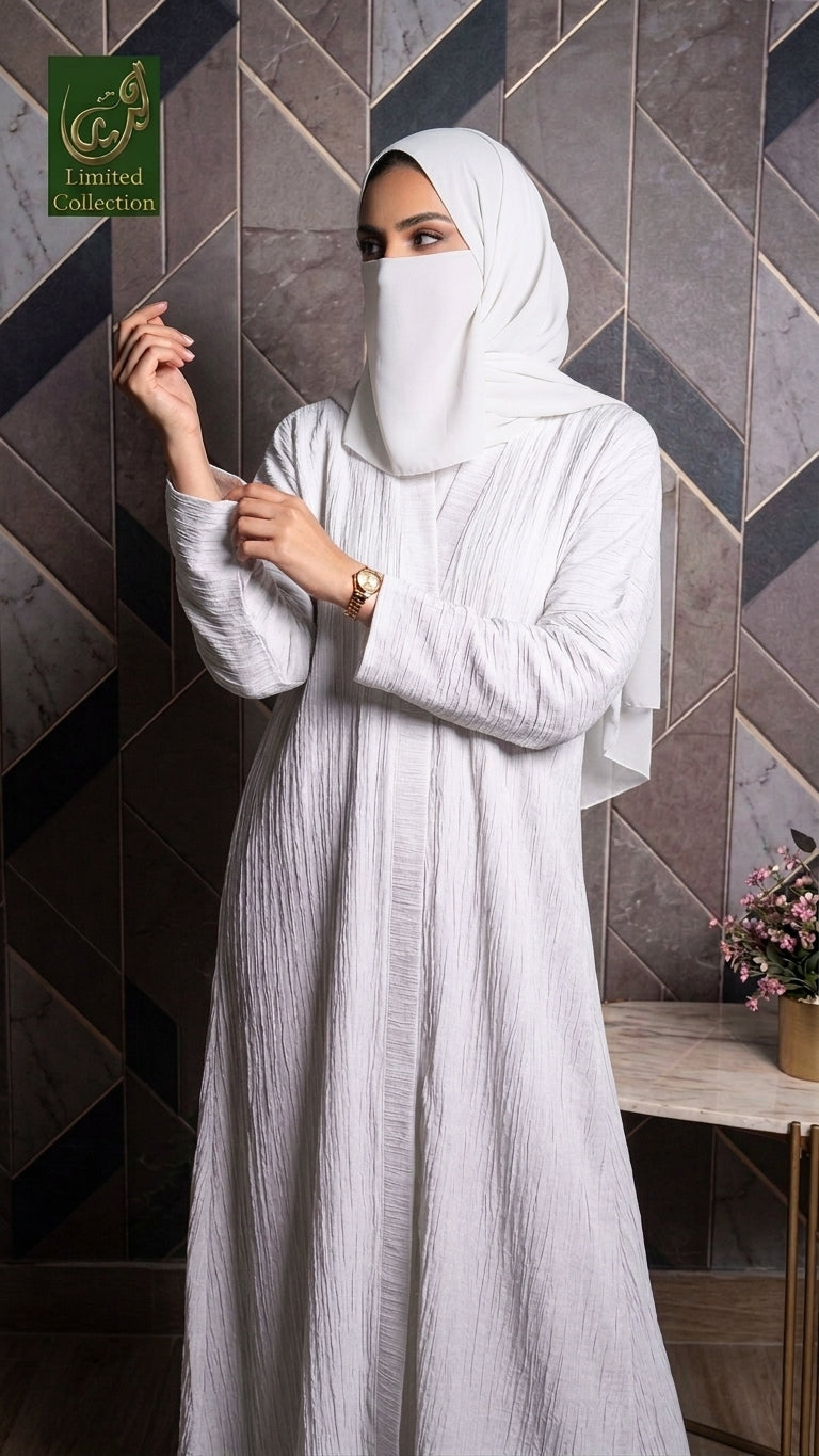 A woman wearing a white textured abaya and matching hijab stands in front of a dark tiled wall. The abaya has long sleeves and a flowing silhouette, while the hijab covers her head and face except for a small side slit.