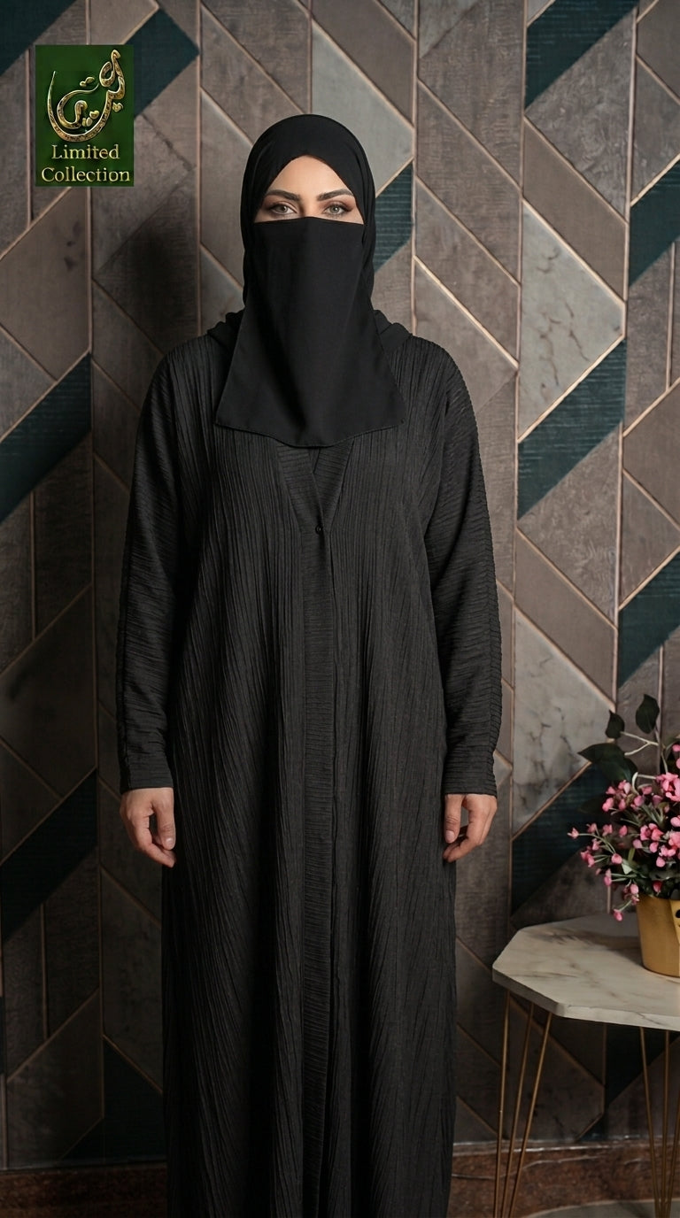 A woman wearing a black long-sleeve abaya and matching black hijab stands against a geometric patterned wall. The abaya has a subtle vertical textured stripe pattern.