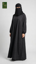 A woman wearing a full-length black abaya and matching black face-covering niqab stands against a light gray studio background. A small green logo appears in the top-left corner.