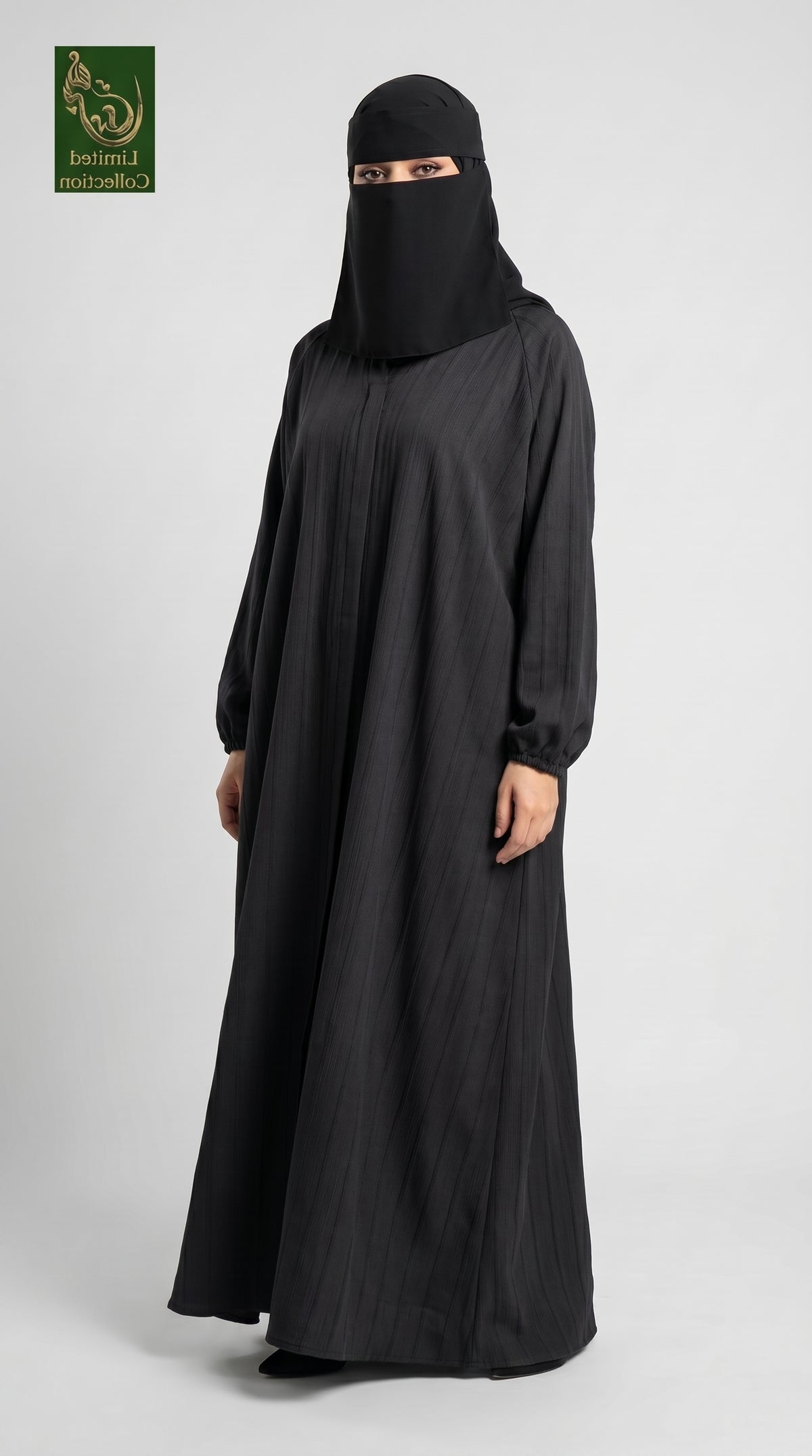 A woman wearing a full-length black abaya and matching black face-covering niqab stands against a light gray studio background. A small green logo appears in the top-left corner.