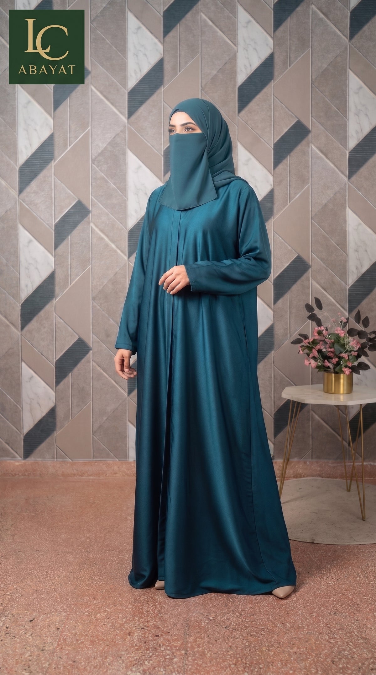 A woman wearing a long teal abaya and matching hijab stands indoors against a geometric patterned wall. The abaya is a deep teal color, and the background features a mix of gray, beige, and brown tiles with a small side table holding a vase of flowers.
