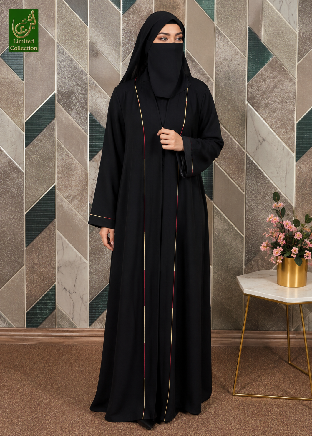 A woman wearing a black Korean Nida Piping work abaya and matching face covering, standing in front of a geometric patterned wall. The abaya has thin gold and red trim details down the front.
