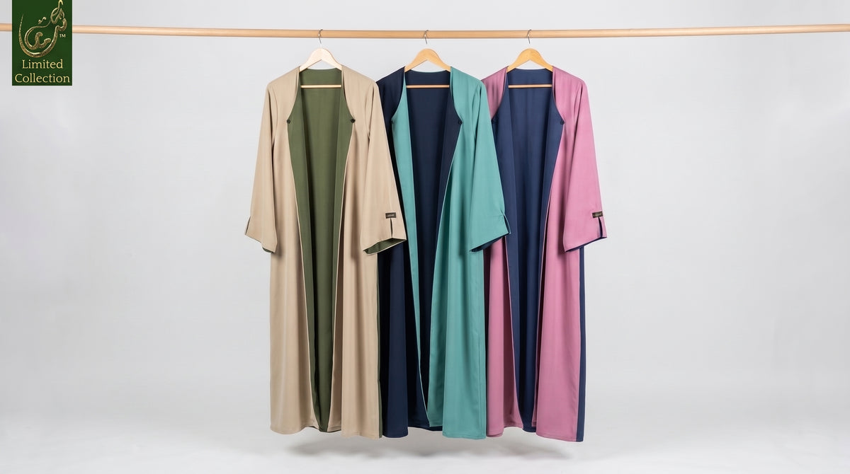 Four long open-front reversible abaya hanging on wooden hangers from a horizontal rod against a light gray studio background. The garments are shown in four colorways: beige over olive green, teal over navy, and pink and purple over navy.