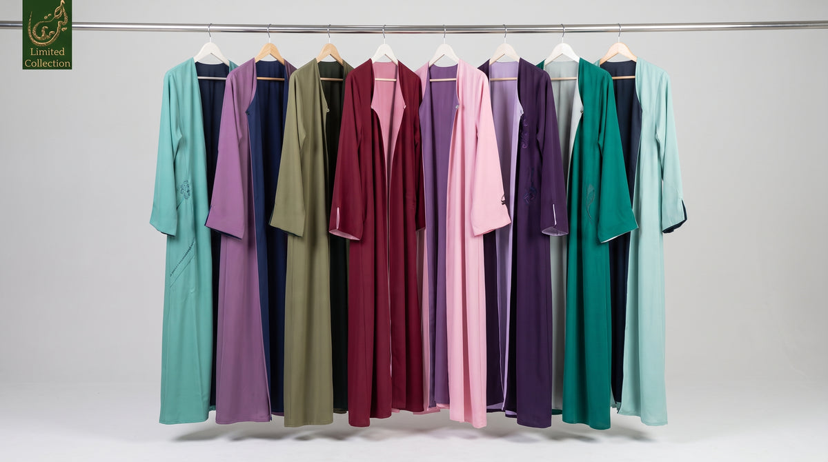 A collection of long, loose-fitting open-front-open reversible abayas in multiple colors, hanging on white hangers from a metal rod against a light gray studio background. The garments include teal, lavender, navy, olive green, burgundy, pink, light purple, gray, dark teal, and mint green.