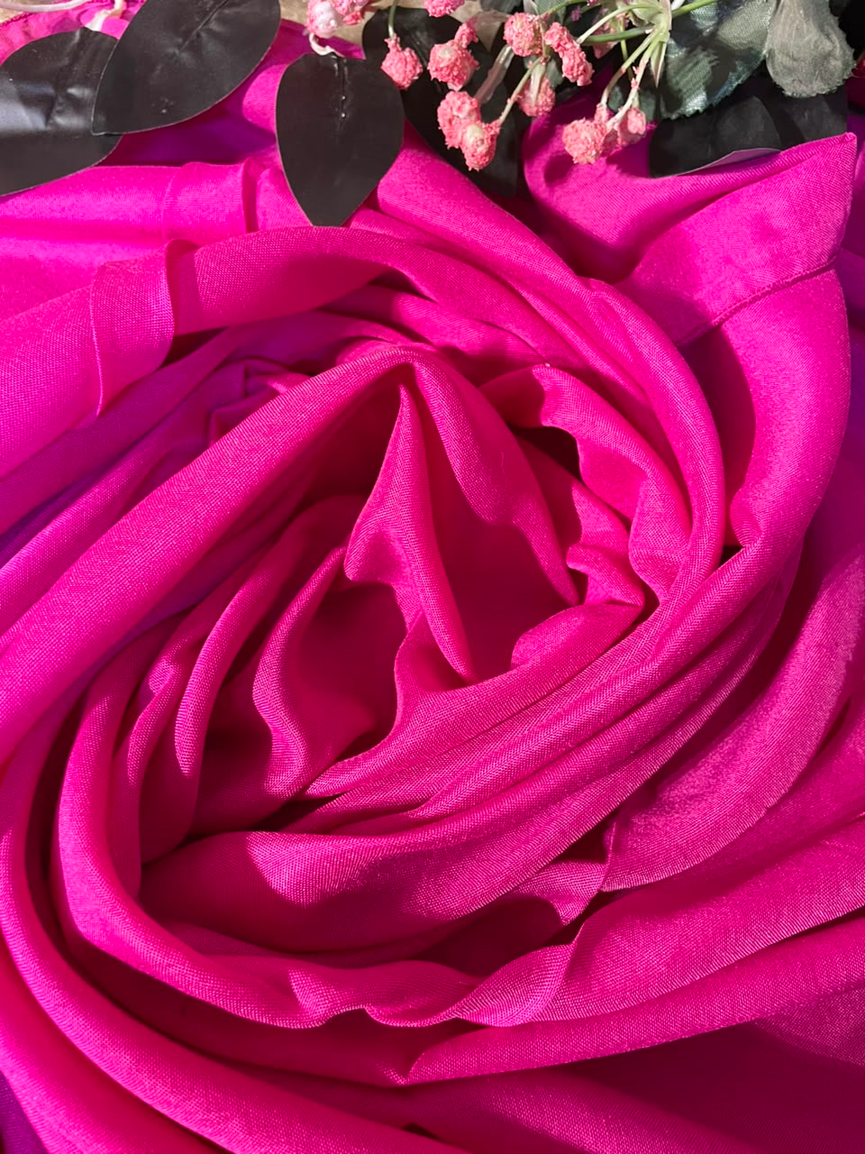 A close-up of draped bright pink fabric arranged in a rose-like spiral, with small dried pink flowers and dark leaves placed at the top edge.