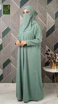 A woman wearing a sage green abaya and matching hijab stands in front of a geometric tiled wall. The abaya is a long, loose-fitting dress with a front opening, and the hijab covers her head and face except for a small lower face opening.