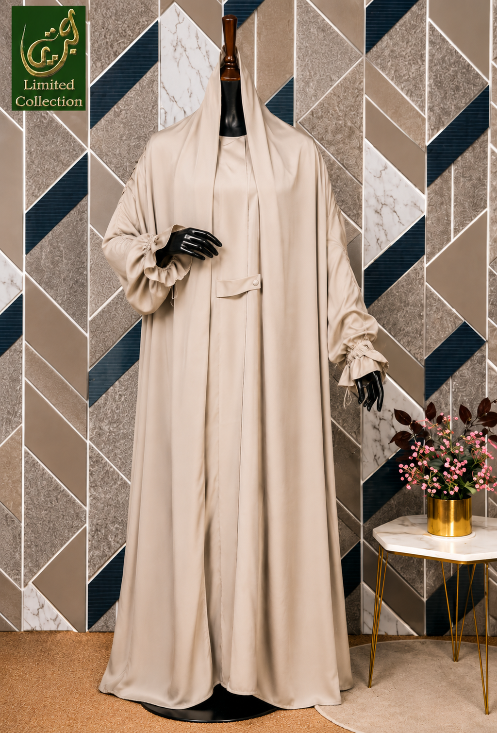 A beige long jilbab dress displayed on a black mannequin set against a geometric patterned wall with a small side table holding flowers. A green 'Limited Collection' logo is visible in the top left corner.