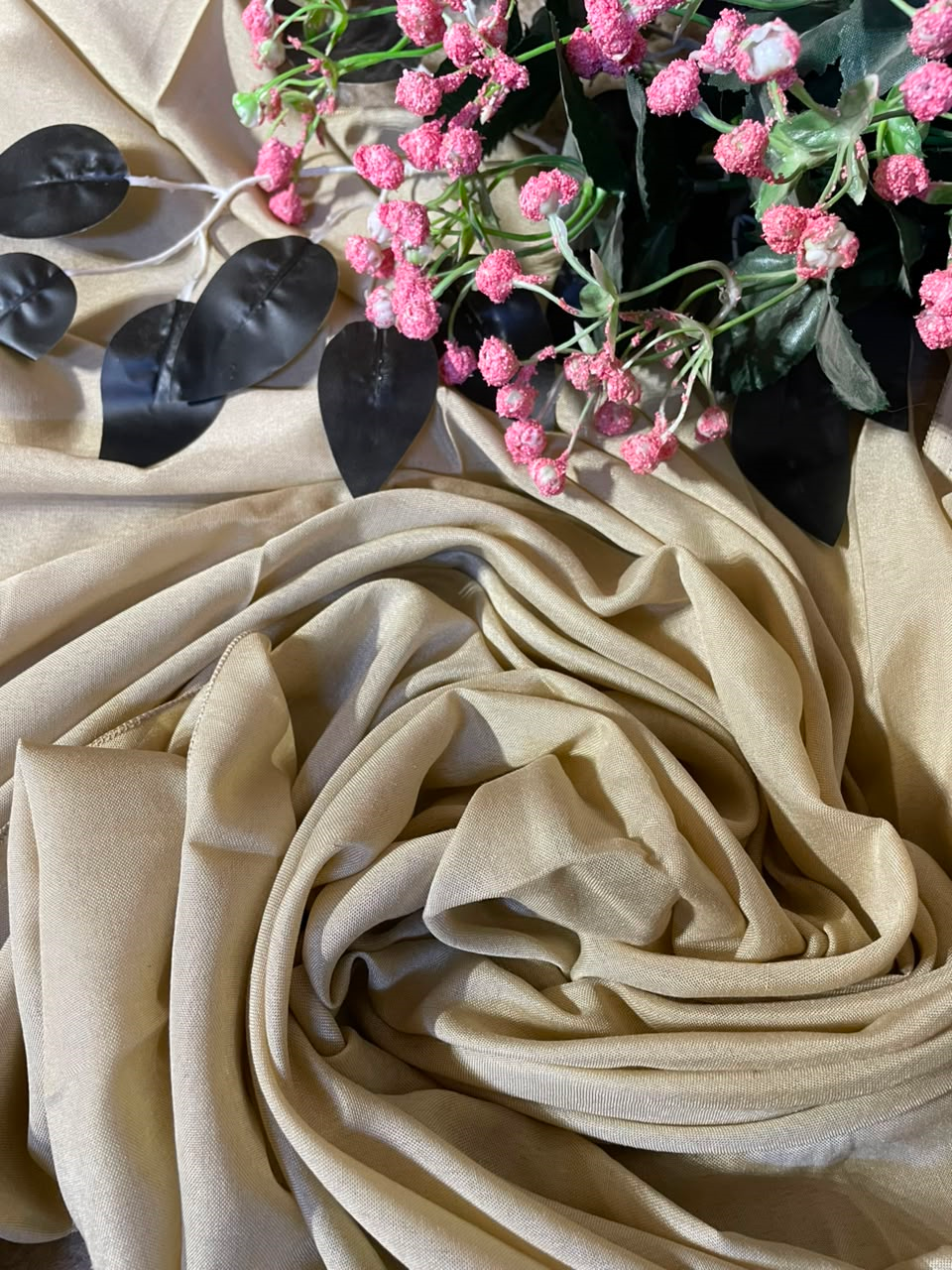 A draped beige fabric scarf or shawl arranged in soft folds, styled with pink flowers and dark leaves on a matching beige fabric background.