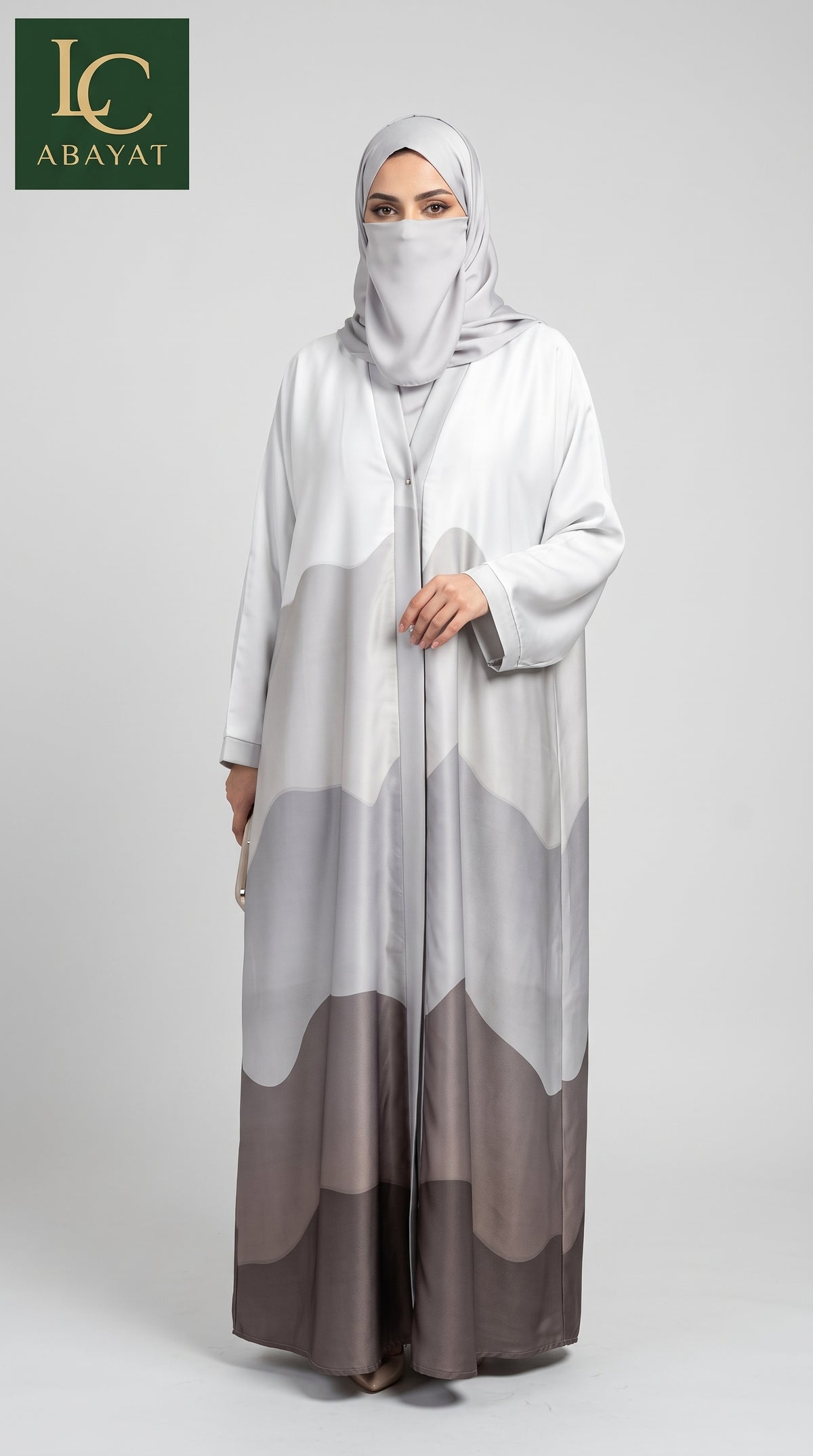 A woman modeling a long, flowing abaya with a matching hijab. The abaya features a mountain-like abstract pattern in shades of white, light gray, and taupe-brown.