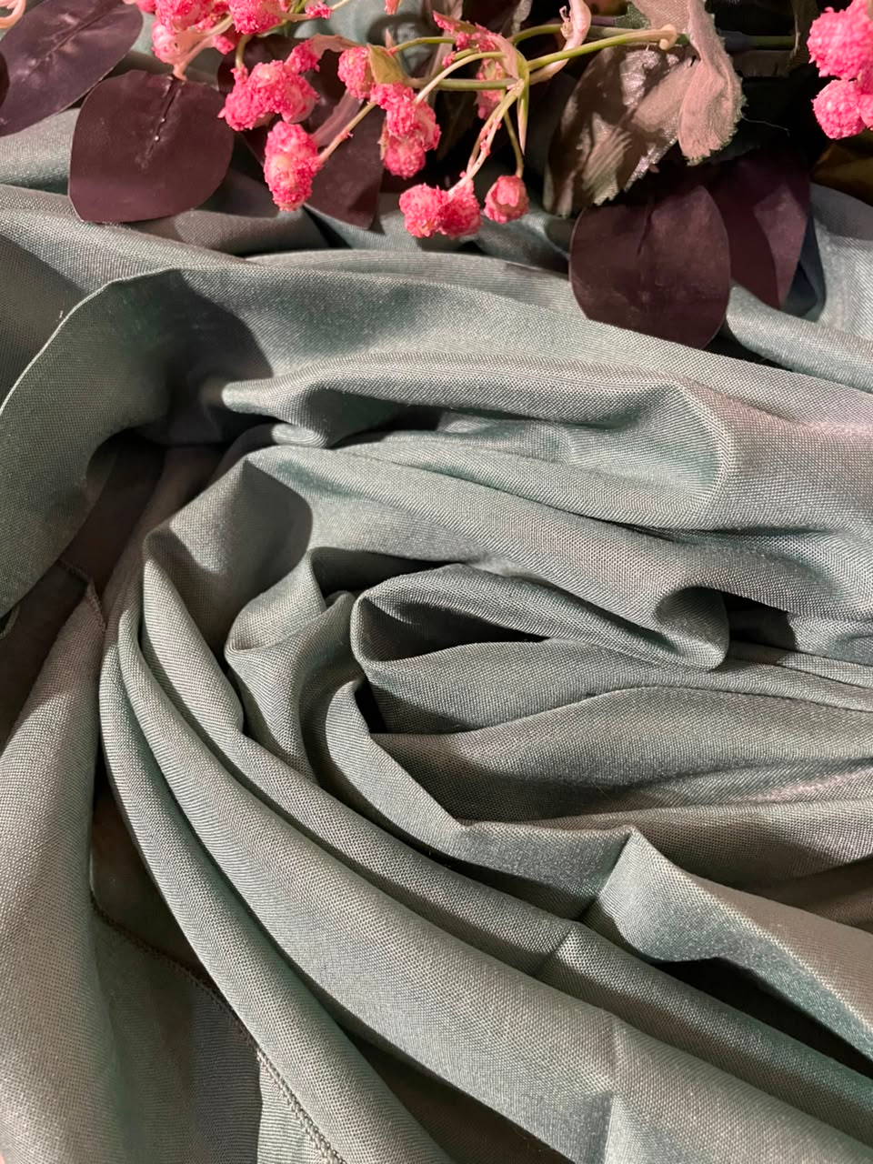 A close-up of draped sage green fabric with soft folds, styled with pink flowers and dark leaves at the top edge.