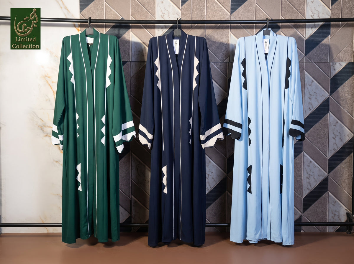 Three long open-front cutwork Abayas hang on a rack against a geometric tiled wall. The left robe is emerald green with white geometric accents, the center robe is dark navy with white geometric accents, and the right robe is light blue with black geometric accents.