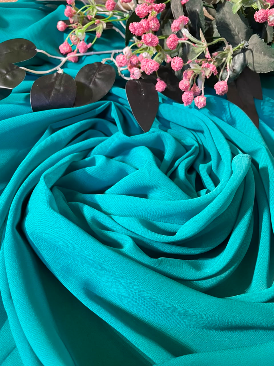 A draped piece of sky blue-green fabric arranged in soft folds, styled with pink flowers and dark leaves at the top against a matching fabric background.