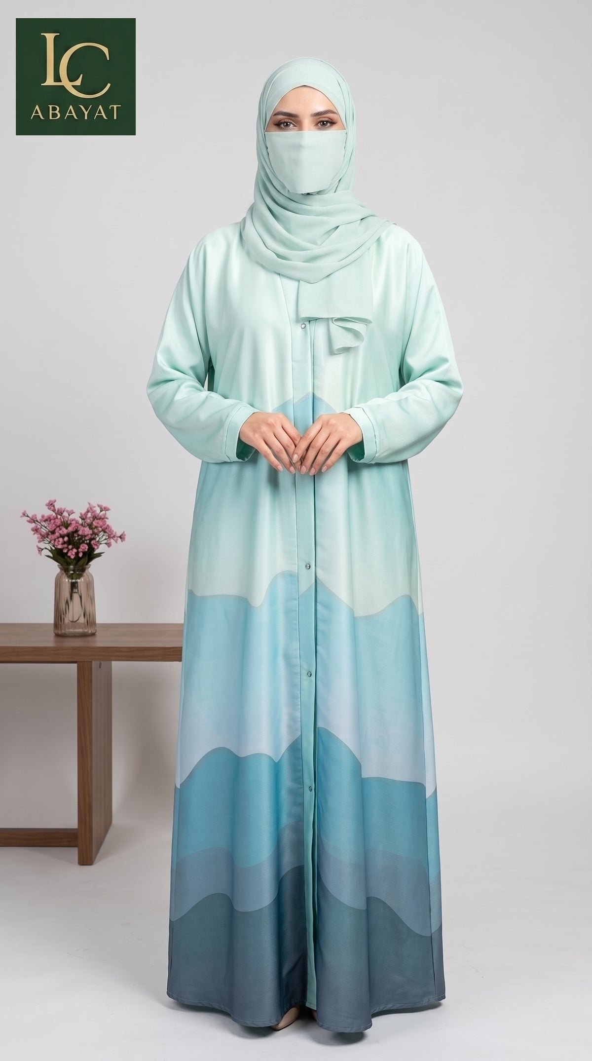 A woman wearing a long mint green and blue gradient abaya with a matching hijab stands in a studio setting. The abaya features a wavy, layered blue pattern at the bottom.