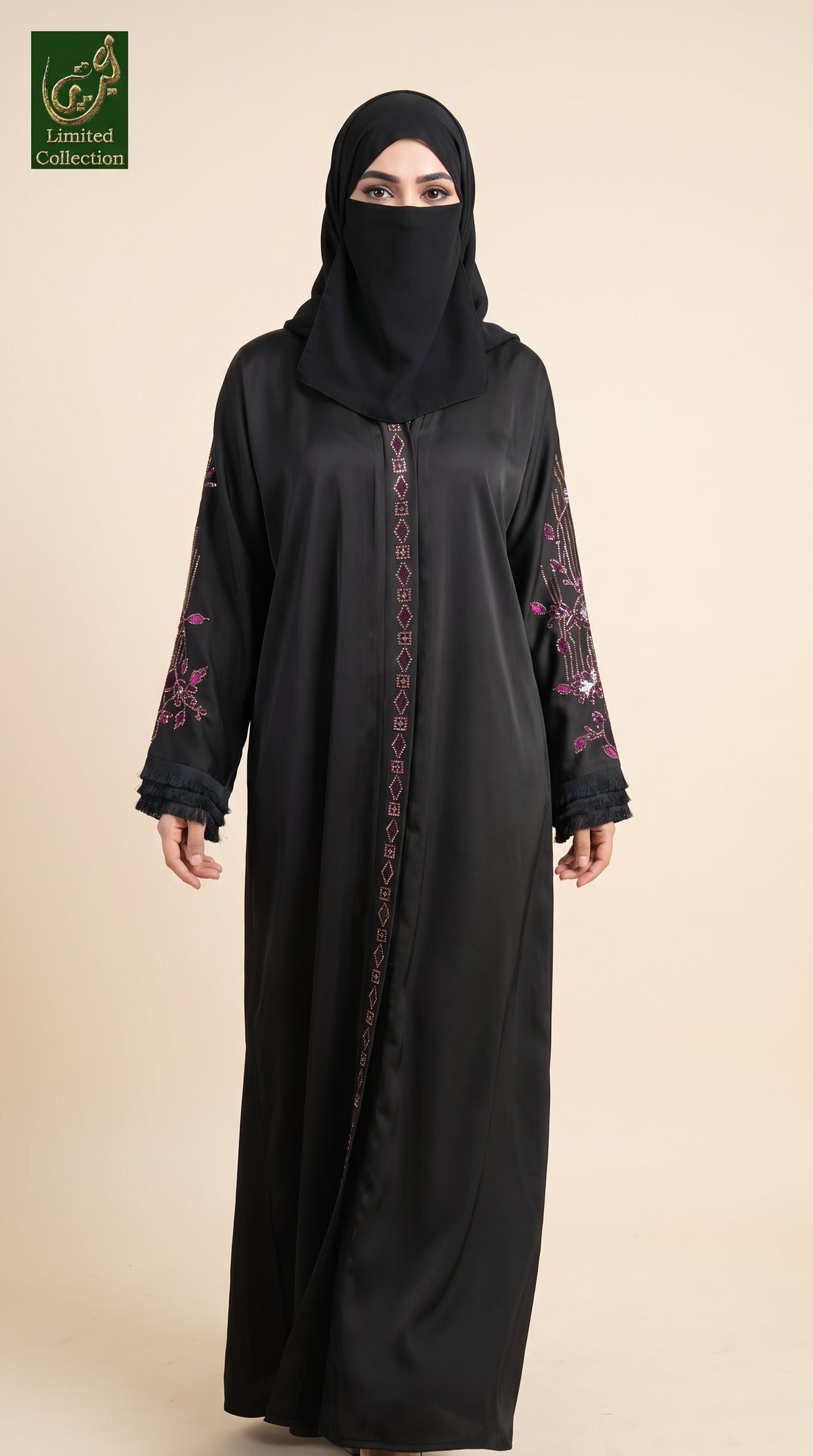A woman wearing a black abaya with pink floral embroidery on the sleeves and a matching black hijab, standing against a solid beige background. A green 'Abayat Limited Collection' logo is visible in the top left corner.