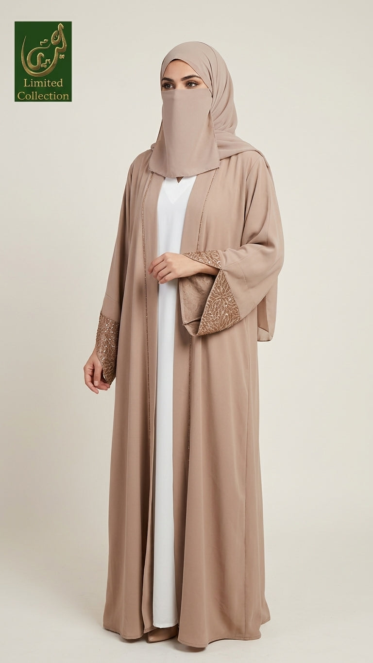 A woman modeling a long beige abaya with wide sleeves and embroidered cuffs, worn over a white inner dress, with a matching beige hijab. The photo is taken against a plain light beige studio background.