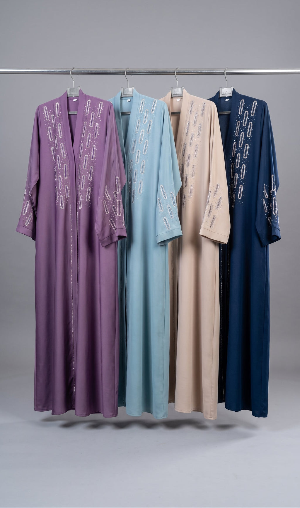 Four long open-front stonework embroidery abayas hang on a metal rack against a light gray studio background. From left to right, the robes are purple, light blue, beige, and navy, each with white embroidered geometric patterns along the front and sleeves.