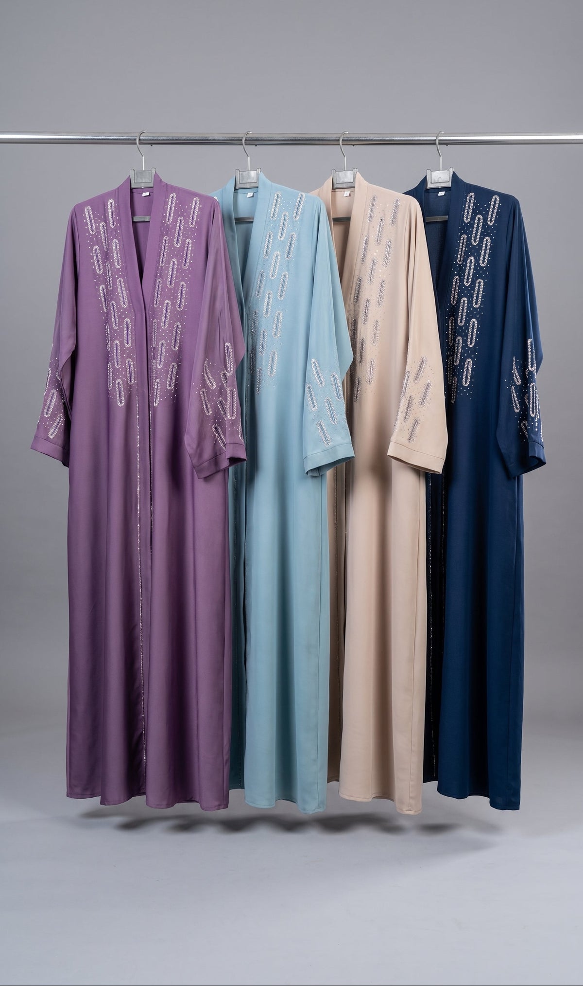 Four long open-front stonework embroidery abayas hang on a metal rack against a light gray studio background. From left to right, the robes are purple, light blue, beige, and navy, each with white embroidered geometric patterns along the front and sleeves.
