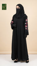 A woman modeling a black abaya with long sleeves featuring red and white floral embroidery, paired with a matching black hijab and black gloves, set against a solid beige background.